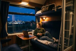 Traveler sleeping in a cozy night train cabin with luggage overhead and city lights passing outside the window