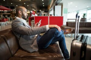 Traveler charging smartphone using portable power bank in airport lounge with AC outlets visible in background