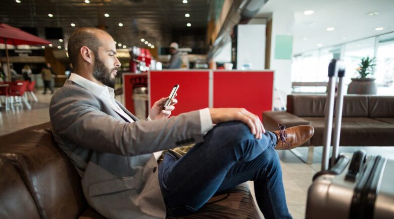 Traveler charging smartphone using portable power bank in airport lounge with AC outlets visible in background
